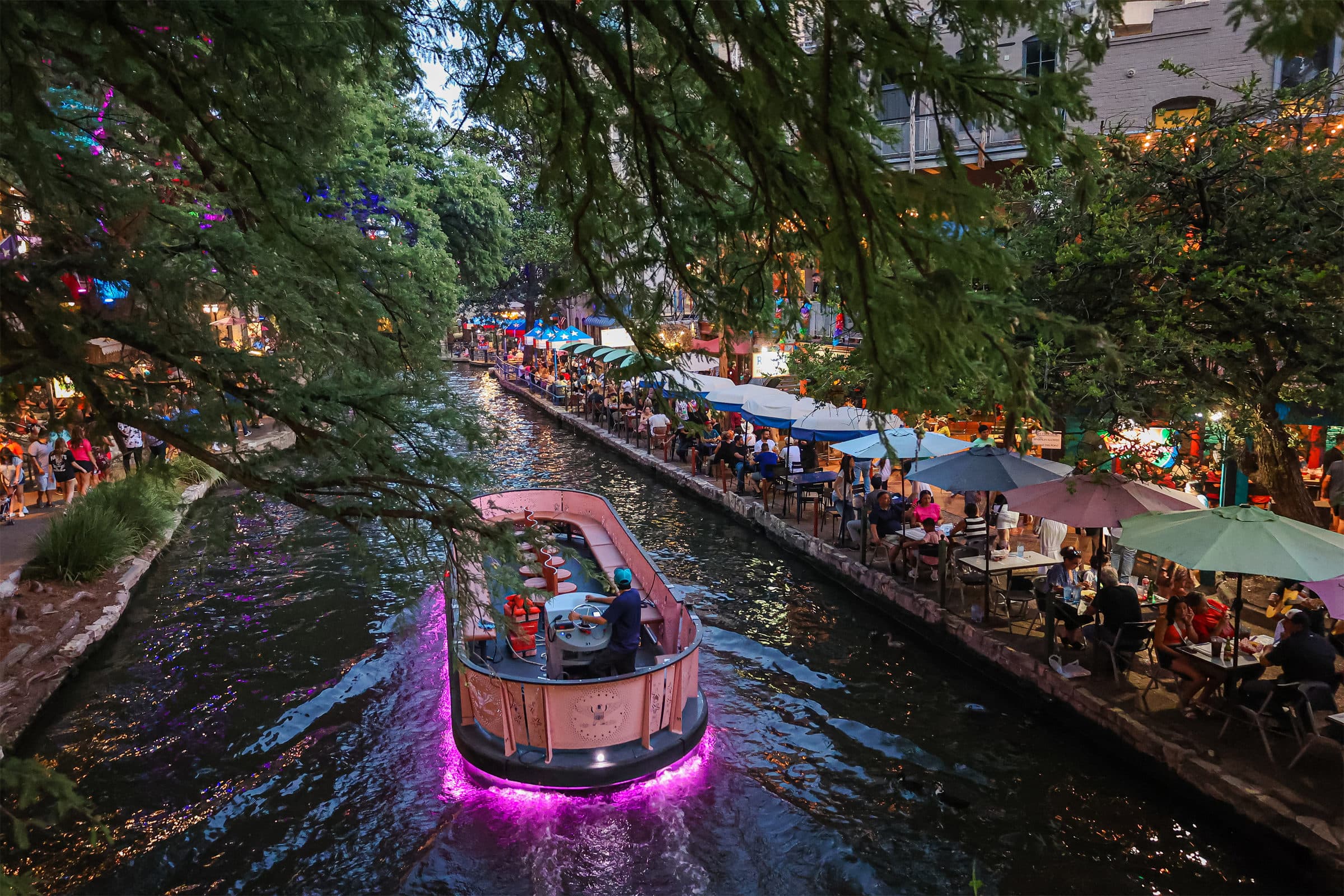 A tour boat on the San Antonio River Walk with riverside dining under the trees